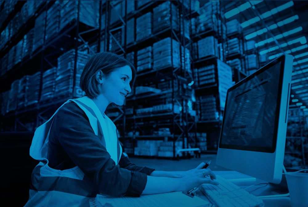 A woman wearing a high-visibility vest is working on a computer in a warehouse filled with tall racks of stacked goods. She is smiling and appears focused while typing and holding a pen. The image has a blue color overlay.